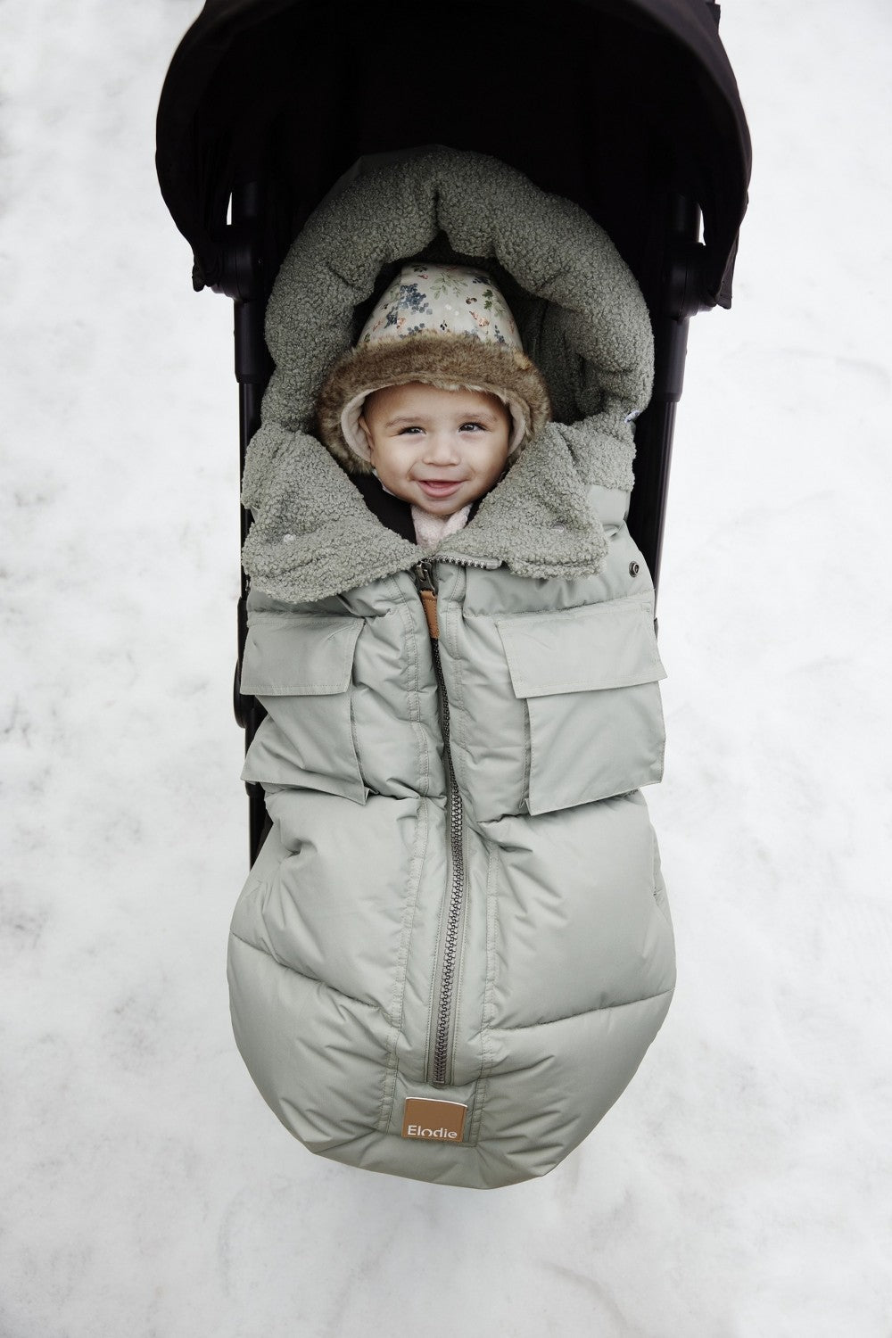 Baby in stroller snuggled in a Hazy Jade footmuff, perfect for winter warmth with thick padding and insulated panels.