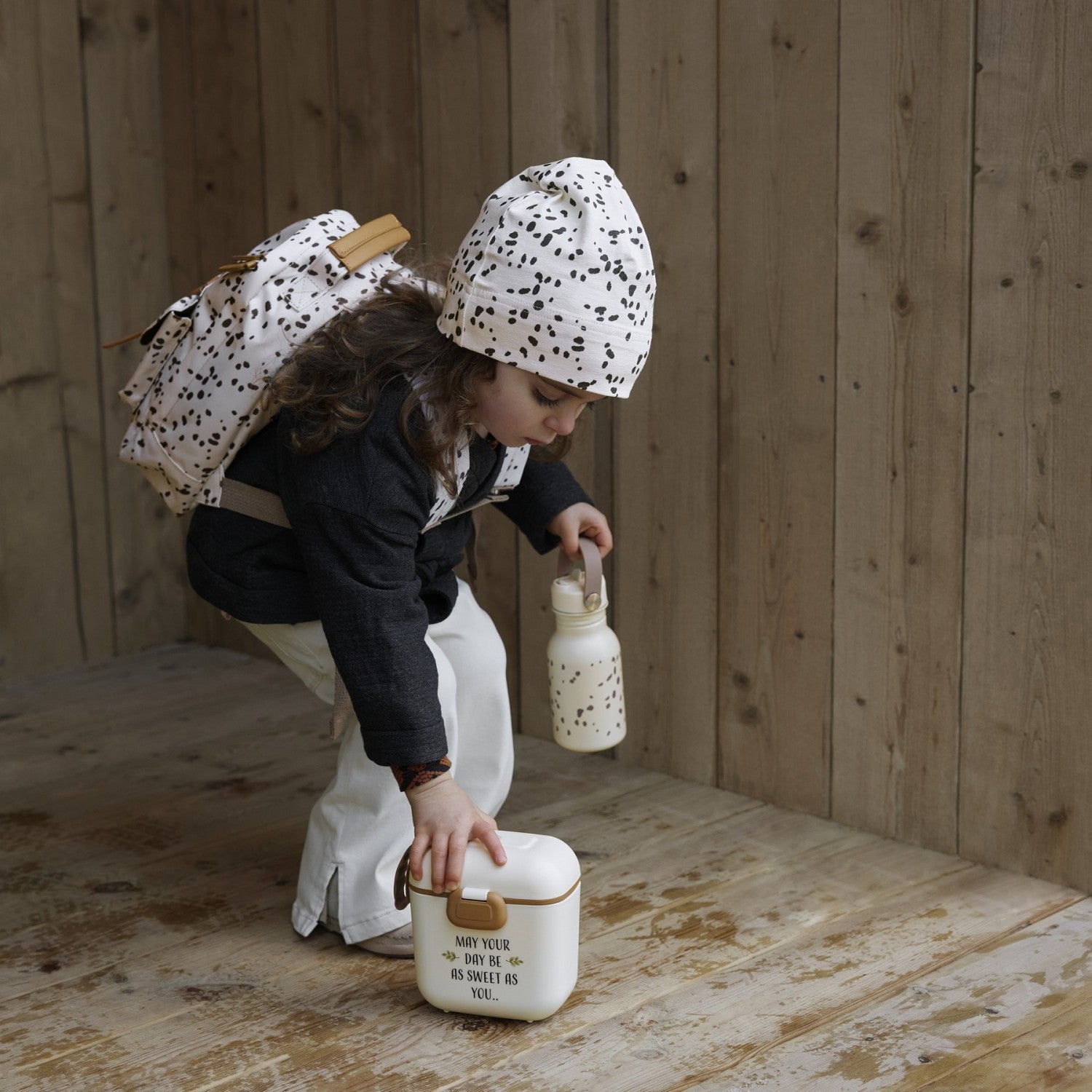 Child wearing Dalmatian Dots kinderrucksack, preparing for an adventure with matching accessories in a rustic wooden setting.