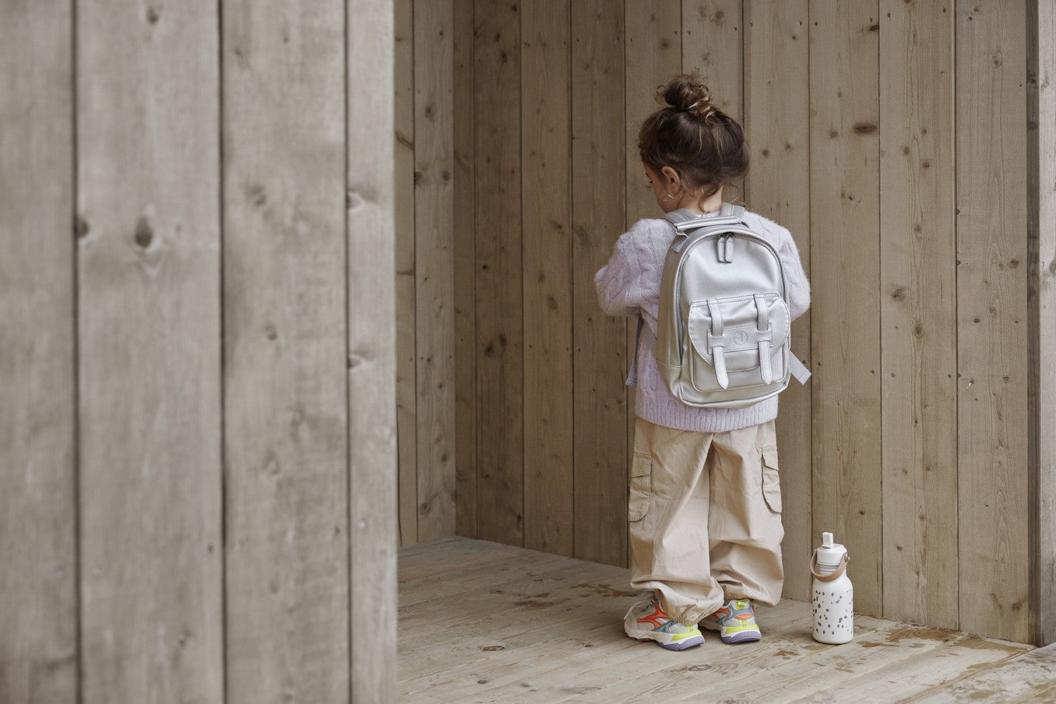 Child wearing Silver Sheen Kinderrucksack exploring wooden cabin with water bottle nearby