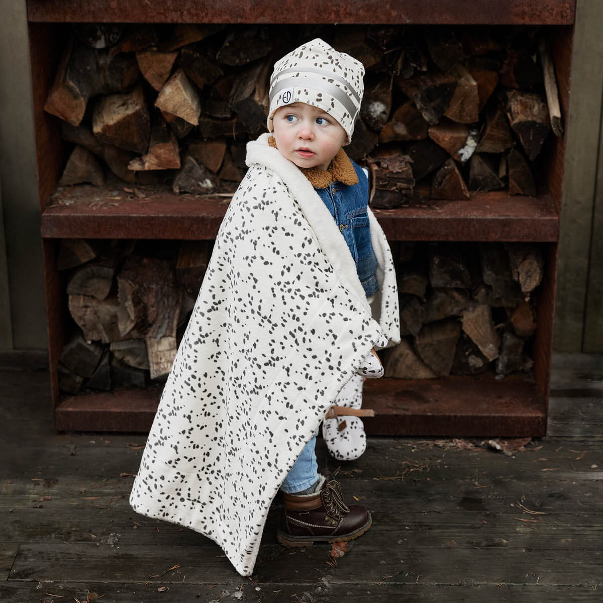 Toddler wearing Dalmatian dots mittens and hat, wrapped in matching blanket, standing in front of log pile.