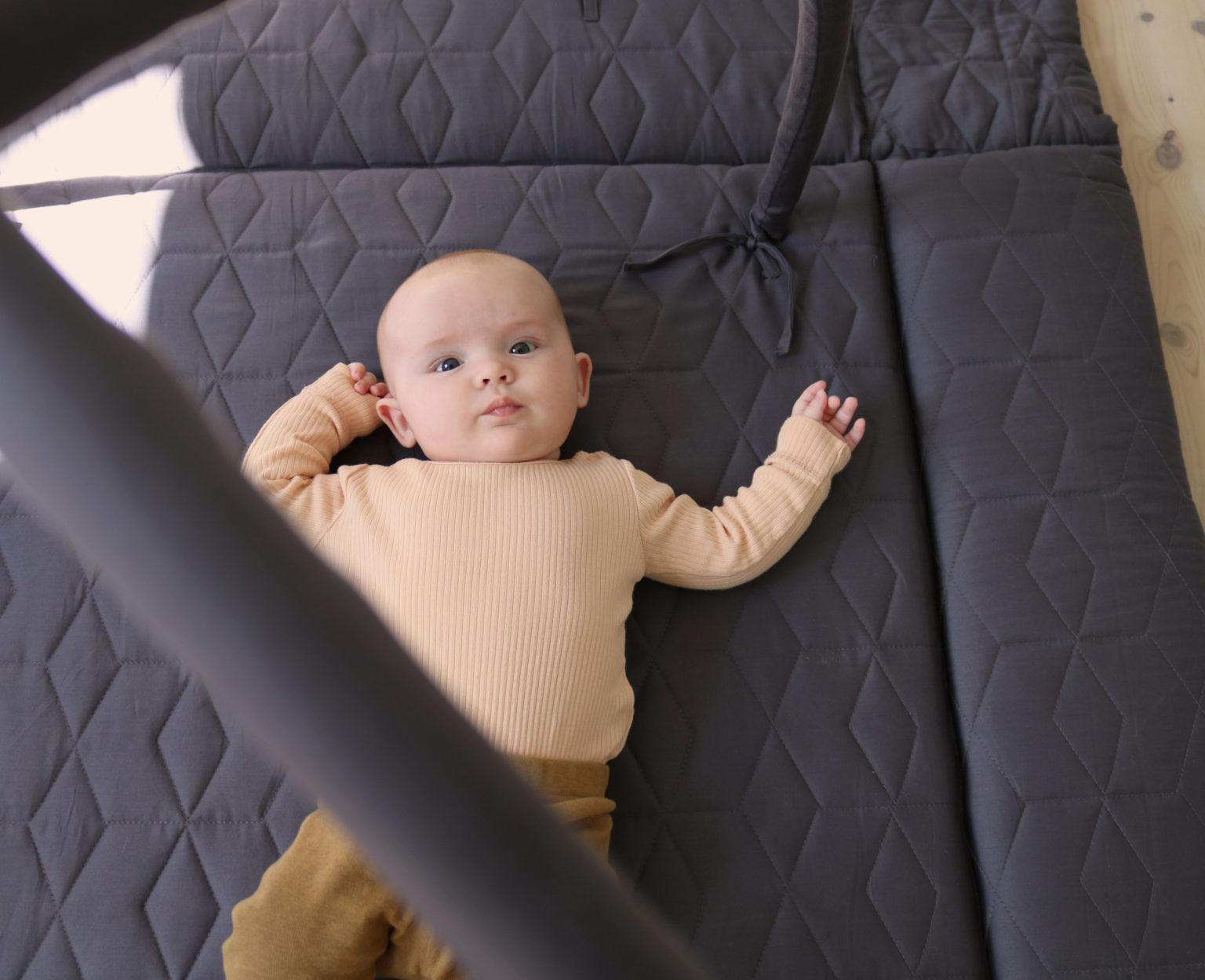 Ein Baby liegt auf einer grauen, gesteppte Spielmatte. Das Baby trägt einen beigen Pullover und braune Hosen und blickt direkt in die Kamera. Der Spielbogen ist teilweise im Bild sichtbar.
